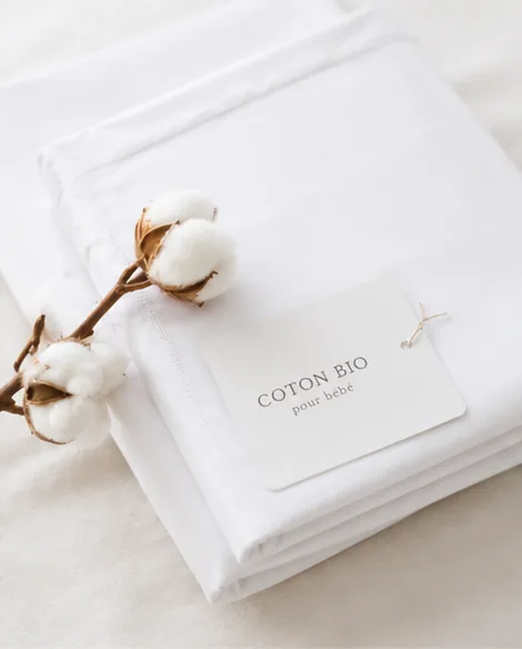Juego de sábanas Cotton Bio de bebé de algodón 100% orgánico de La Cama Azul, doblado junto a una rama de algodón